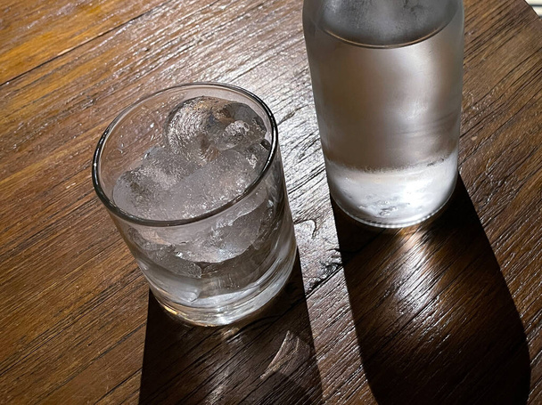 Seçici odak, buzla dolu berrak bir bardak ve temiz bir cam şişe soğuk maden suyunu ahşap masaya koyar, susuzluğu gidermek için su kullanır. - Fotoğraf, Görsel