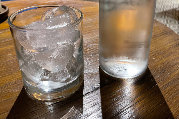 Selektivní zaostření, Čiré sklo naplněné ledem a čiré skleněné láhve drží studenou minerální vodu sedí na dřevěném stole, voda použít k uhašení žízně - Fotografie, Obrázek