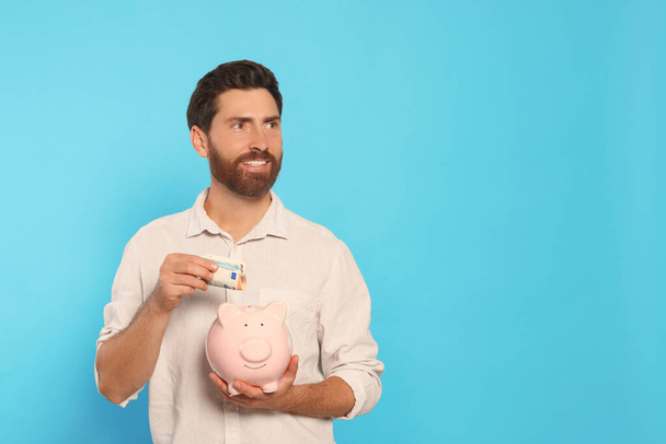 Happy man putting money into piggy bank on light blue background, space for text - 写真・画像