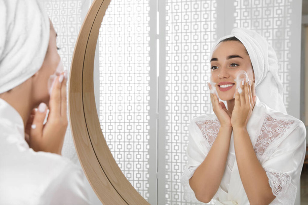 Beautiful young woman applying cleansing foam onto face near mirror in bathroom. Skin care cosmetic - Foto, imagen
