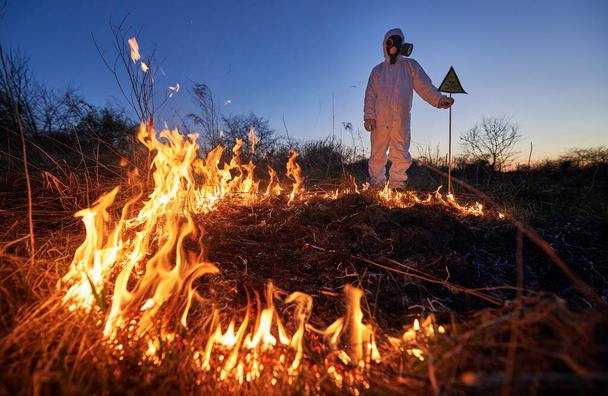 Firefighter ecologist working in field with wildfire at night. Man in suit and gas mask near burning grass with smoke, holding warning sign with skull and crossbones. Natural disaster concept. - Zdjęcie, obraz