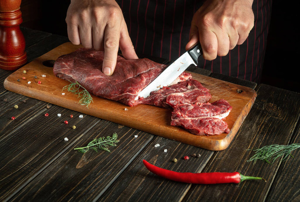 The cook cuts raw fresh beef meat on a cutting board before baking or barbecue. Working environment in the kitchen of a restaurant or hotel - Photo, Image
