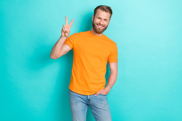 Portrait of confident nice guy with blond beard wear orange t-shirt hand in pocket showing v-sign isolated on teal color background. - Фото, изображение