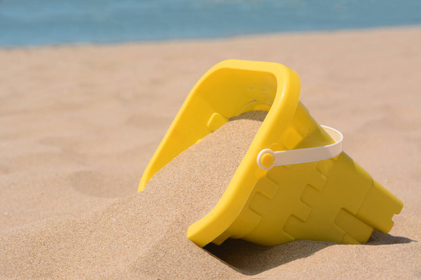 Yellow plastic bucket on sand near sea, closeup. Beach toy - Valokuva, kuva