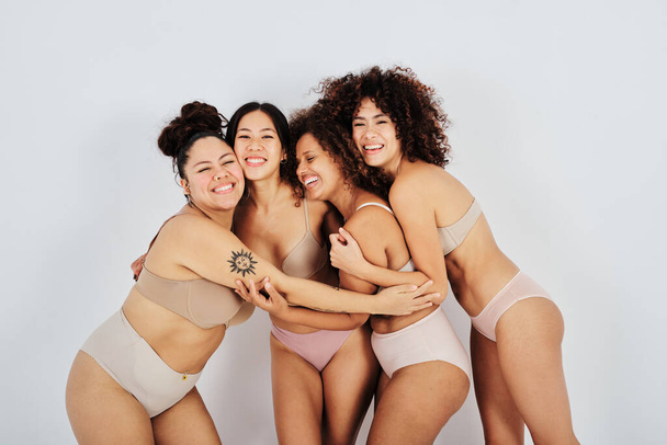 Veselý multiracial ženské kamarádky ve spodním prádle objímání zatímco stojí proti šedé pozadí ve studiu - Fotografie, Obrázek