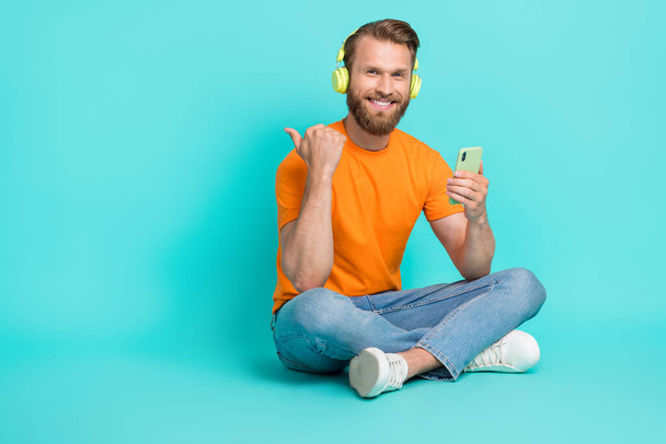 Full size photo of handsome man wear orange t-shirt jeans headphones directing empty space hold phone isolated on teal color background. - Foto, Imagem
