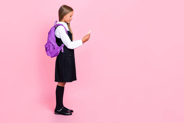 Full length side profile photo of clever positive girl with blond hair wear uniform look at smartphone isolated on pink color background. - Photo, Image