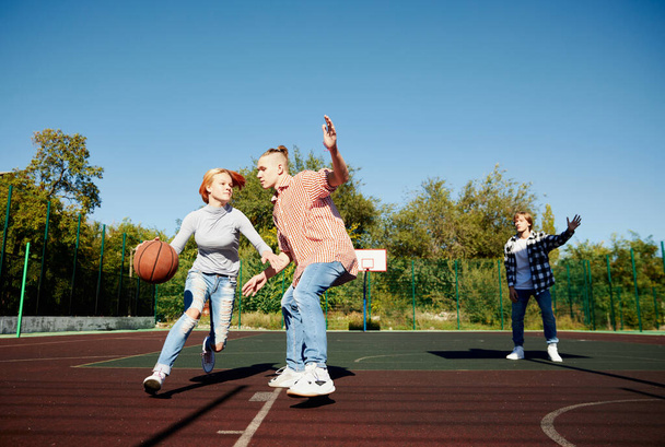 Group of teens, students playing street basketball at basketball court outdoors at spring sunny day. Sport, leisure activities, hobbies, team, friendship. Boys and girl spending time together. - Фото, изображение