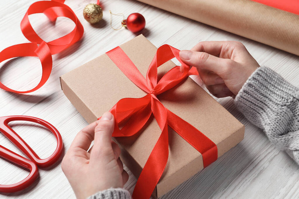Weihnachtsgeschenk. Frau bindet Schleife auf Geschenkbox an weißem Holztisch, Nahaufnahme - Foto, Bild