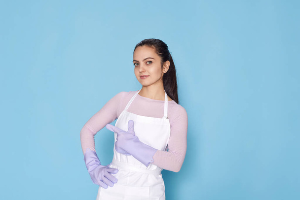 beautiful caucasian woman in violet rubber gloves pointing fingers aside at copy space on blue background. cleaning - Фото, изображение