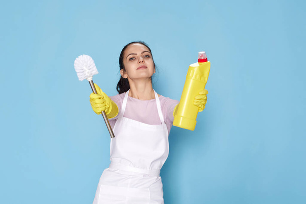 woman in yellow rubber gloves and cleaner apron showing toilet brush and bottle of detergent on blue background. - Foto, Imagem