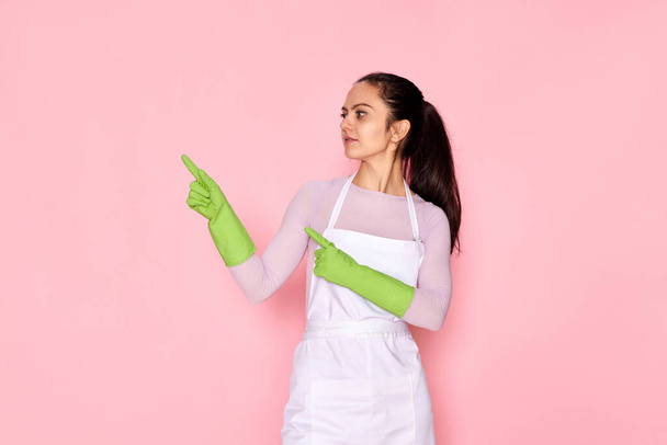 beautiful caucasian woman in rubber gloves pointing fingers aside at copy space on pink background. cleaning - Photo, image