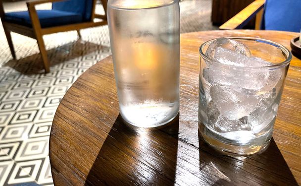 Enfoque selectivo, un vidrio transparente lleno de hielo y botella de vidrio transparente tiene agua mineral fría se sienta en la mesa de madera, uso de agua para saciar la sed - Foto, Imagen