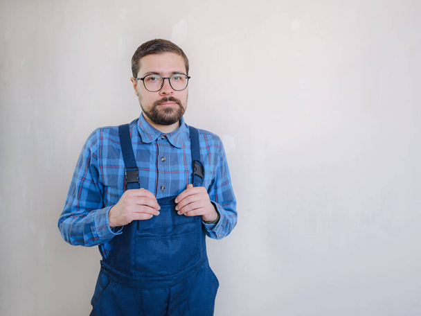 young man in blue work suit looking at camera on white wall background. Repair apartment. Home renovation concept. - Foto, afbeelding