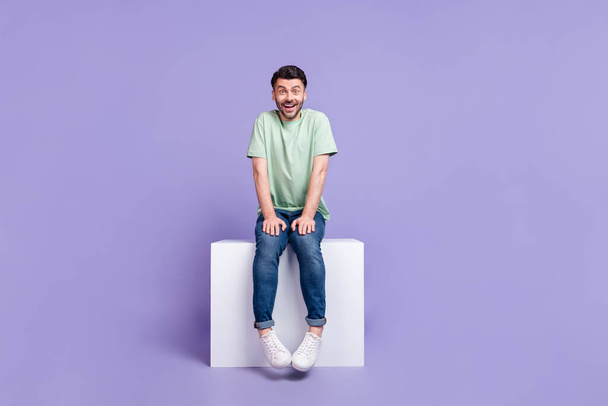Full body photo of attractive young man sitting white podium shy excited dressed stylish gray garment isolated on purple color background. - Fotó, kép