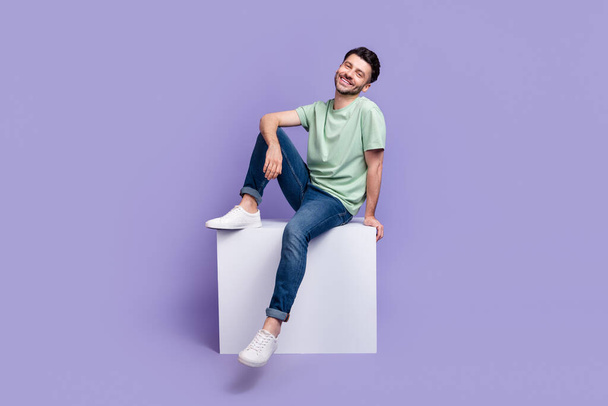 Full length photo of nice young man sitting white copyspace cube cheerful dressed stylish gray clothes isolated on purple color background. - Fotografie, Obrázek