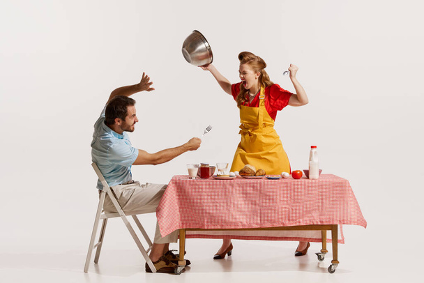 Portrait of young emotive couple loudly, aggressively quarreling while doing breakfast isolated on white background. Concept of retro style, domestic duties, old-fashion, lifestyle. Copy space for ad - Foto, Imagen