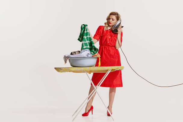 Portrait of beautiful woman standing with bowl filled with clothes and talking on iron like on phone. Concept of retro style, domestic duties, old-fashion, lifestyle. Copy space for ad - Photo, Image