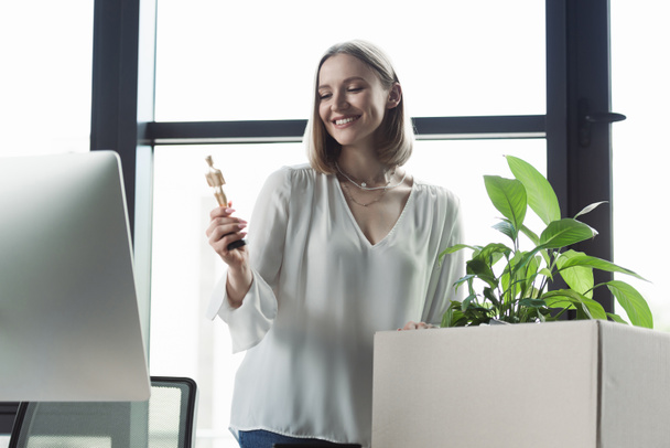Cheerful intern holding doll near carton box with plant and computer in office  - Photo, Image