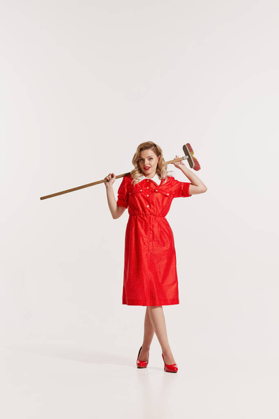 Portrait of beautiful woman in styles red dress posing with mop isolated over white background. Concept of retro style, domestic duties, old-fashion, lifestyle. Copy space for ad - Фото, изображение