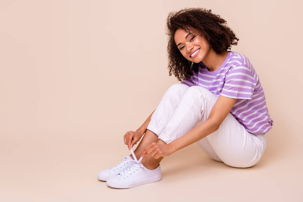 Portrait of optimistic nice cute girl wavy hairstyle wear striped t-shirt white trousers sneakers sit isolated on beige color background. - Фото, зображення