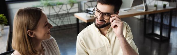 Glimlachende manager die brillen aanraakt terwijl hij praat met stagiaire in kantoor, spandoek  - Foto, afbeelding