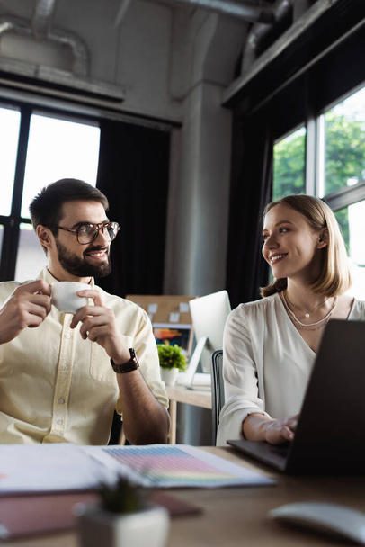 Веселый стажер смотрит на бизнесмена с чашкой кофе возле ноутбука и бумаги в офисе  - Фото, изображение