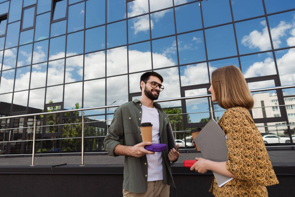 Hombre de negocios sonriente sosteniendo lonchera y café para acercarse a colega con carpeta de papel al aire libre  - Foto, Imagen