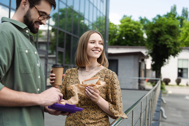 Femme d'affaires souriante tenant un sandwich et un café à emporter près d'un collègue dans une rue urbaine  - Photo, image