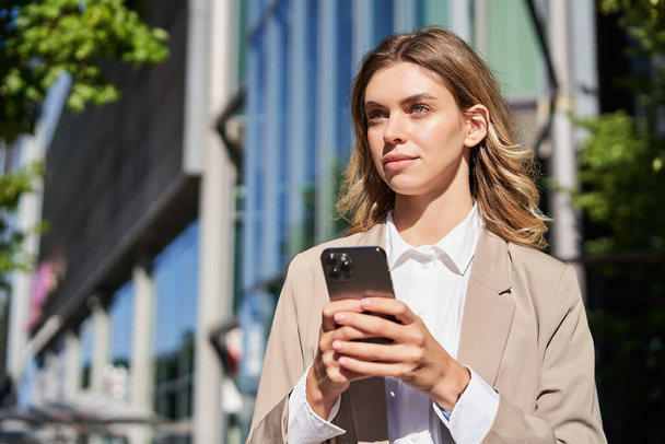 Портрет стильной деловой женщины в корпоративной одежде, держащей смартфон, стоящей на улице и пользующейся мобильным телефоном в руках. - Фото, изображение