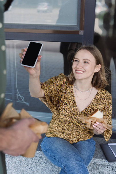 Mujer de negocios sonriente sosteniendo sándwich y teléfono inteligente cerca de colega en la calle urbana  - Foto, imagen