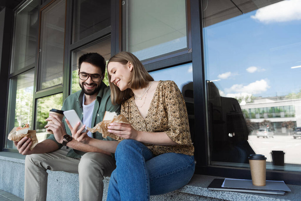 Empresária sorridente mostrando smartphone para colega com sanduíche e café para ir ao ar livre  - Foto, Imagem