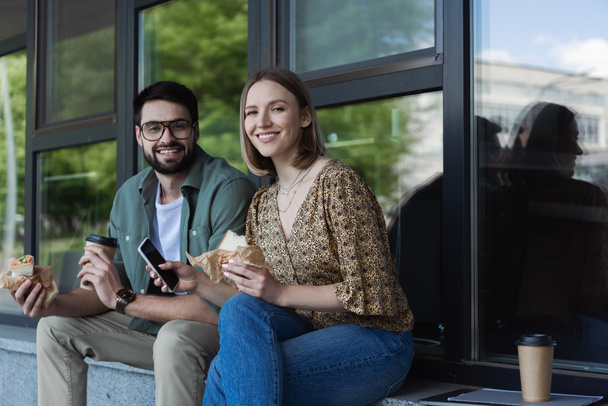 Gente de negocios sonriente sosteniendo sándwiches y teléfonos inteligentes mientras mira la cámara cerca del edificio al aire libre  - Foto, imagen