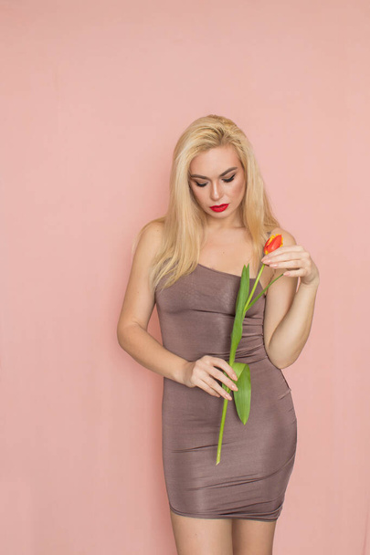 Jonge mooie zelfverzekerde vrouw in trendy zomeravond bruine jurk. Sexy zorgeloze blonde vrouw poseren in de buurt van roze muur in de studio. Model met rode lippen. Geïsoleerd. Een rode tulp in haar handen - Foto, afbeelding