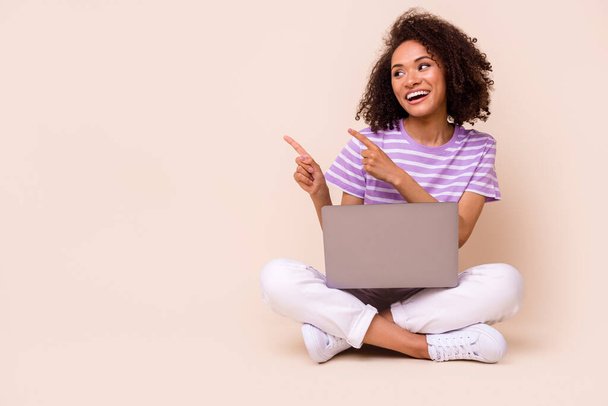 Photo of cheerful lovely girl brunette hair wear striped t-shirt directing look empty space sit laptop isolated on beige color background. - 写真・画像
