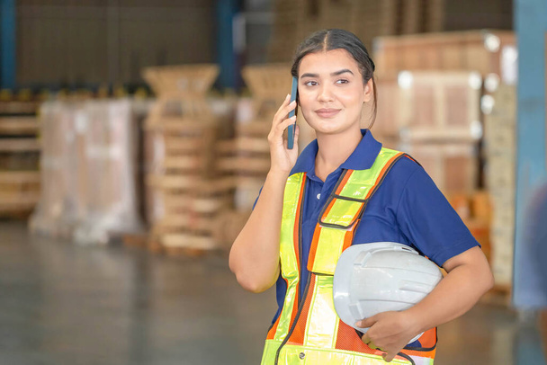 Νεαρή γυναίκα αποθηκάριος στέκεται στην αποθήκη με τη χρήση κινητού smartphone, χαμογελώντας γυναίκα σε σκληρό καπέλο, εργαζόμενος που εργάζεται στη λογιστική βιομηχανία μέσα σε αποθήκη εργοστάσιο - Φωτογραφία, εικόνα
