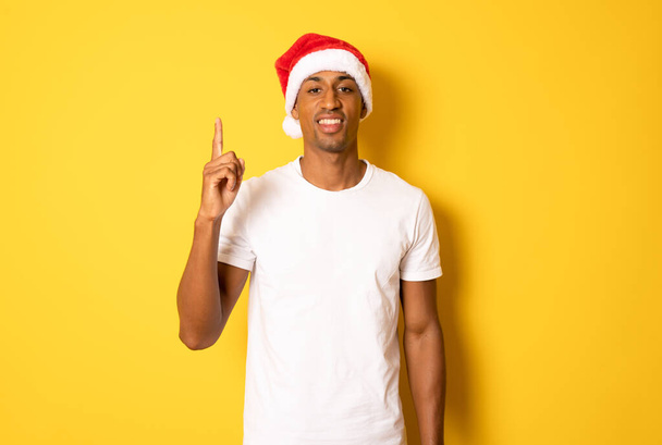 Jonge Afro-Amerikaanse man met tevreden uitdrukking wijzende vinger omhoog, draagt Santa hoed, feliciteert u met Nieuwjaar, draagt casual jumper, geïsoleerd over gele achtergrond. De Kerstman komt naar je toe! Vakantie concept - Foto, afbeelding