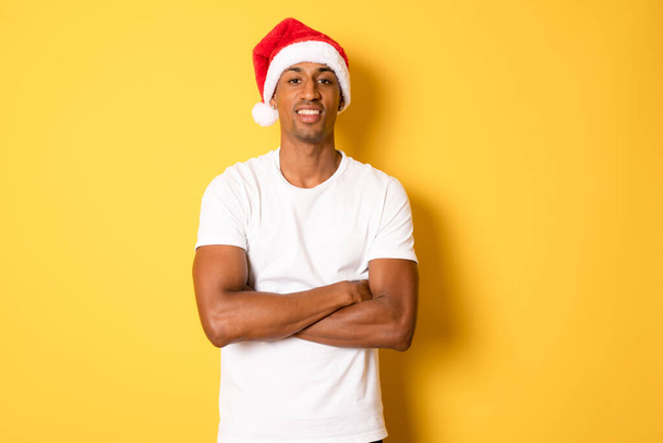 Nuori onnellinen afrikkalainen amerikkalainen mies jouluhattu pystyssä kädet taitettuna eristetty keltaisella pohjalla. - Valokuva, kuva