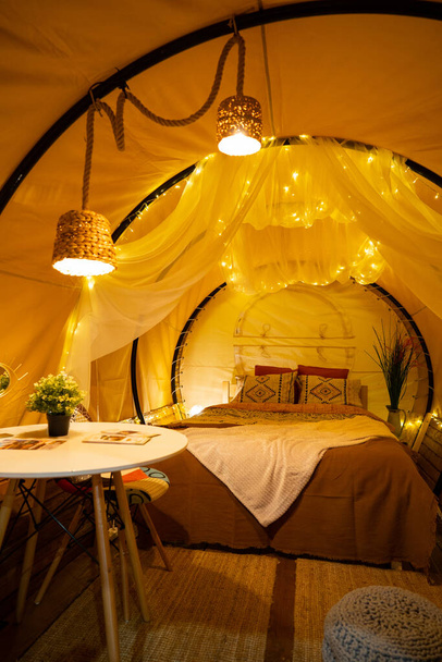 Color image from inside a tent, with very cozy bed, blankets, pillows and lights, at a glamping. - Photo, Image