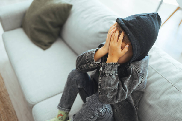 Depressed sad child sitting on the bed, at home. The little boy is hiding his head between hands. Upset problem child with head in hands, concept for childhood bullying, depression stress or frustration - 写真・画像
