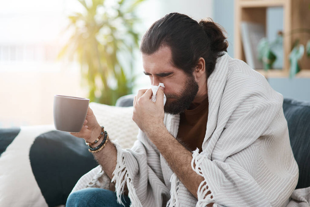 Zieke dagen zijn het ergst. een zieke jongeman die zijn neus snuit met een zakdoekje in zijn woonkamer - Foto, afbeelding