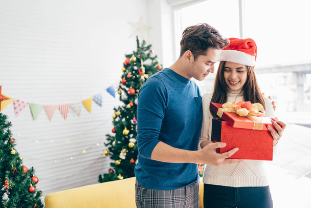 サンタクロースの帽子をかぶっている若い美しい幸せなアジアの女性は、背景にクリスマスツリーがある家でクリスマスプレゼントで彼女のボーイフレンドを驚かせます。コピースペース付き画像. - 写真・画像