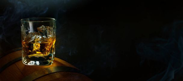 A glass of whiskey with ice on an oak barrel and the smoke of a Cuban cigar on a dark background. Men's club banner idea. Copy space for text - 写真・画像