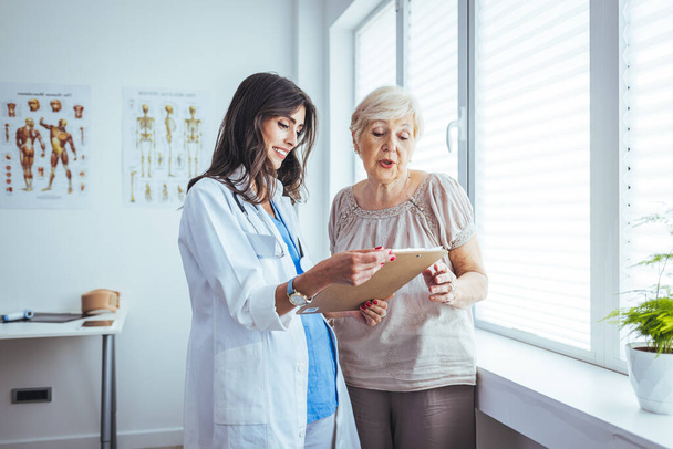 Happy female doctor and senior patient look at clipboard discuss treatment or therapy on gadget. Smiling woman nurse and mature client use pad device discussing results in modern hospital. - Foto, Imagem