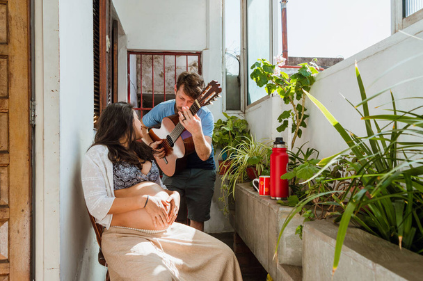young latin couple of bearded caucasian argentinian man singing and playing guitar with his pregnant brazilian woman wife, at home - Foto, Imagen