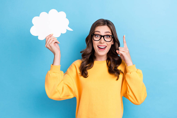 Photo of young funky adorable woman wear yellow shirt toothy smile wear glasses hold paper cloud finger up idea decision isolated on blue color background. - Zdjęcie, obraz
