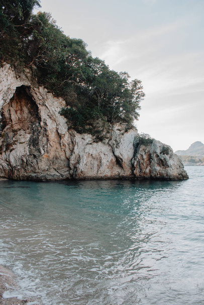 Corfú, Grecia. Una enorme roca en la playa con agua azul en la que crecen plantas y árboles verdes. - Foto, imagen