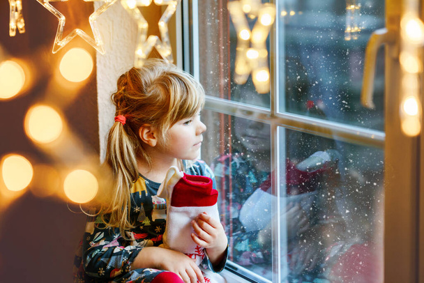 Niña preescolar sosteniendo la bota de Santa Claus taza con regalo llamado Nikolausstiefel en alemán. Niño feliz espera en vacaciones por ventana con luces de Navidad en invierno. Acogedora celebración familiar de Navidad - Foto, imagen