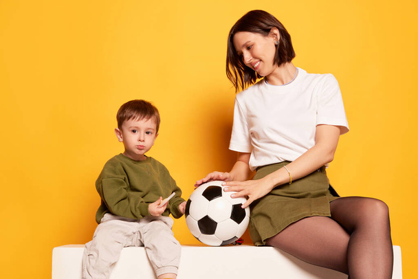 Χαμογελώντας νεαρή μητέρα με τον χαριτωμένο μικρό γιο της κρατώντας μπάλα ποδοσφαίρου απομονωμένη σε φωτεινό κίτρινο φόντο. Οικογένεια, αγάπη, μητρότητα και η έννοια Ημέρα Μητέρας. Φλάιερ με χώρο αντιγραφής για διαφήμιση - Φωτογραφία, εικόνα