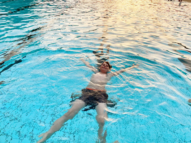 Школьный мальчик плескается в открытом бассейне в теплый летний день. Счастливый здоровый ребенок дошкольного возраста наслаждается солнечной погодой в городском общественном бассейне. Активность детей на открытом воздухе с водой - Фото, изображение